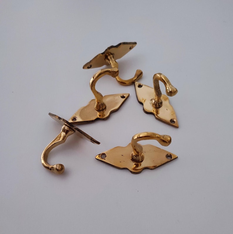 May include: A collection of five antique brass hooks. Each hook has a curved design and is mounted on a decorative backplate. The hooks are in various shapes, including diamond and oval, and are ideal for hanging items.