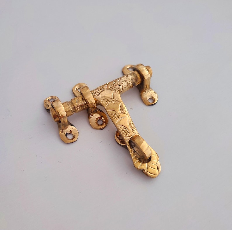 May include: A golden, ornate door latch with a floral design. The metal latch has a T-shape with a sliding bolt. The latch is designed to be mounted on a door frame. The measurements are in centimetres.