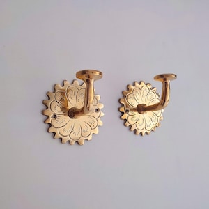 Könnte beinhalten: Zwei Wandhaken aus Messing. Jeder Haken hat eine kreisförmige, floral gemusterte Rückplatte mit einem gebogenen Haken, der sich von der Mitte aus erstreckt. Die Haken sind goldfarben und das Design ist mit geätzten Linien detailliert.
