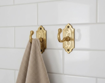 May include: Two ornate brass wall hooks with a floral design are mounted on a white tiled wall. One hook holds a beige towel. The hooks have a curved design and are secured with small screws, adding a touch of vintage charm to the bathroom.