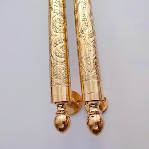 May include: Two ornate, gold-colored scroll holders with intricate floral and swirl patterns. Each holder has a cylindrical body and a rounded, bulbous base. The holders are mounted on round, gold-colored wall plates.