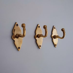 May include: Three brass wall hooks. Each hook has a decorative, diamond-shaped backplate with a curved hook. The hooks are a shiny gold color and are mounted on a light gray wall.