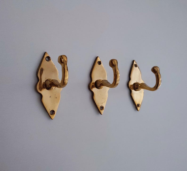 May include: Three brass wall hooks. Each hook has a decorative, diamond-shaped backplate with a curved hook. The hooks are a shiny gold color and are mounted on a light gray wall.