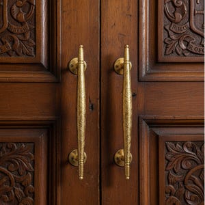 May include: A pair of decorative, brass-coloured door handles on a dark wooden door. The handles have an intricate pattern and are mounted on circular plates. The door panels are made of dark wood and feature carved floral designs.