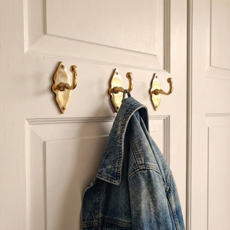 May include: Three antique brass coat hooks mounted on a white door. The hooks have a decorative, ornate design and are holding a denim jacket. The hooks are approximately 4 inches long.