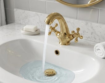 Op de afbeelding: Een goudkleurige badkamerkraan met ingewikkelde bloemendetails. Water stroomt in een witte wastafel. De wastafel heeft een gouden afvoerstop. Een stuk zeep ligt op een klein schaaltje. De kraan is klassiek.