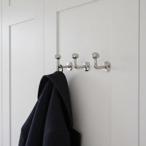 Könnte beinhalten: Drei silberfarbene Kleiderhaken an einer weißen Tür. Ein dunkelblauer Mantel hängt am ersten Haken. Die Haken haben ein abgerundetes Design und sind gleichmäßig verteilt.