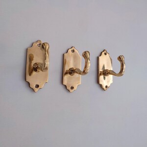 May include: Three brass wall hooks with a decorative design. Each hook has a curved arm and is mounted on a rectangular backplate with a scalloped top edge. The hooks are a warm gold color.