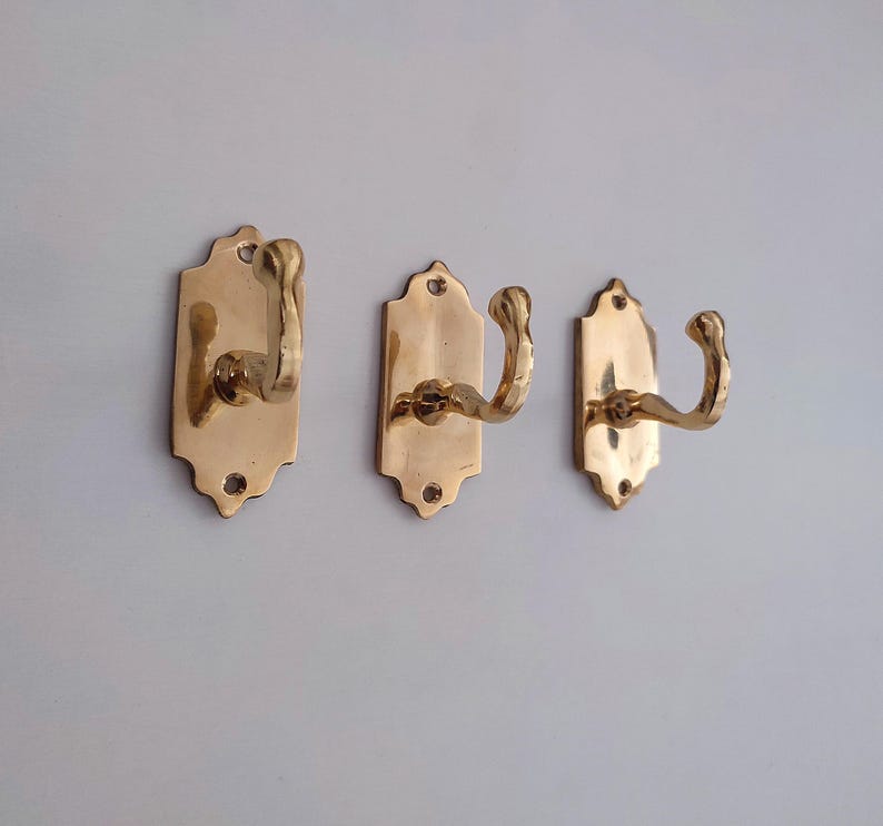 May include: Three brass wall hooks with a decorative design. Each hook has a curved arm and is mounted on a rectangular backplate with a scalloped top edge. The hooks are a warm gold color.