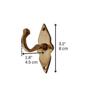 May include: A brass-colored wall hook with a decorative diamond-shaped backplate. The hook is 1.8 inches (4.5 cm) wide and 3.1 inches (8 cm) tall. It has a curved hook design and two screw holes for mounting. The hook is made of metal.