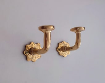 May include: A pair of gold-coloured wall hooks. Each hook features a curved arm and a floral-shaped base. The hooks are mounted on a white wall, highlighting their decorative design. The hooks are made of metal.