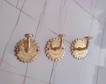 May include: Three brass wall hooks with a decorative, sunburst-like design. Each hook has a curved arm extending from a circular base with etched patterns. The hooks are mounted on a white and grey tiled surface.