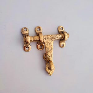 May include: A brass door latch with floral engravings. The latch has a rectangular bar with three screw holes on each end and a decorative handle. The metal has a polished, golden finish.