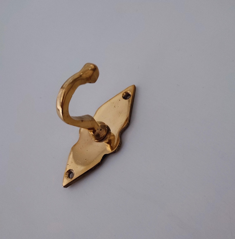 May include: A brass-colored wall hook with a decorative, diamond-shaped backplate. The hook curves upwards, and the backplate has two screw holes for mounting. The metal has a polished finish, reflecting light.