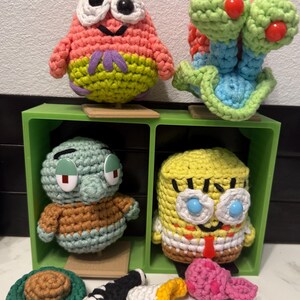 May include: Crocheted figures of cartoon characters, including a pink starfish, a green snail, a green squid, and a yellow sponge. The figures are displayed in a green shelf unit, with additional crocheted accessories.