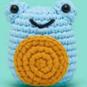 May include: A handmade crochet frog in light blue with a yellow spiral design on its belly. The frog has black eyes and a smiling mouth. The background is a solid light green color.