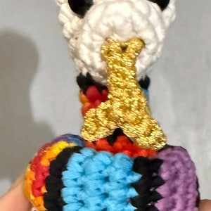 Può includere: Una figura fatta a mano all'uncinetto a forma di uccello, con testa bianca, occhi neri e becco dorato. Il corpo è un patchwork di colori vivaci tra cui blu, arancione, rosso, nero e viola. L'oggetto artigianale è probabilmente un pezzo decorativo.