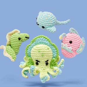 May include: Four handmade crochet sea creatures: a green seahorse, a light blue turtle, a pink fish, and a green octopus. Each creature has black safety eyes and is made with soft yarn. The background is a solid light blue.