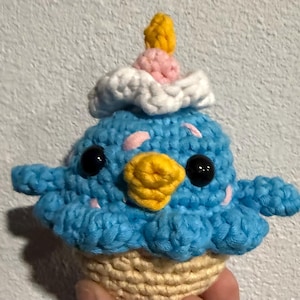 May include: A handmade crochet cupcake featuring a blue bird design. The bird has a yellow beak, black eyes, and pink accents. The cupcake has white frosting, a pink layer, and a yellow candle. The base is beige.
