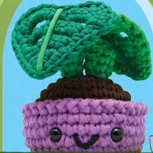 May include: A handmade crochet plant with a smiling face. The plant pot is purple, the soil is brown, and the leaves are green. The plant has black button eyes and a stitched black smile.