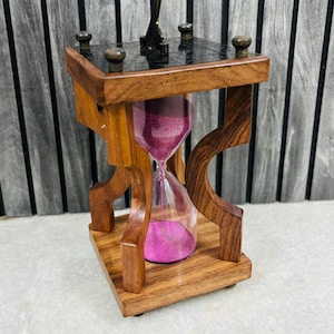 May include: A wooden hourglass with a dark top and base, featuring a clear glass chamber filled with pink sand. The hourglass is supported by a decorative wooden frame with curved legs and a small sundial on top.
