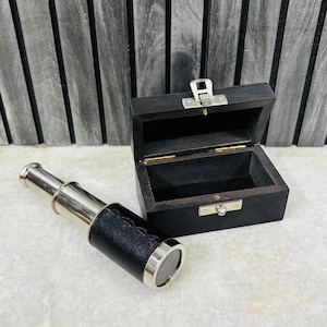 Op de afbeelding: Een vintage-stijl messing en zwarte telescoop met een leren handgreep, naast een open zwarte houten kist. De telescoop heeft een zilverkleurige metalen afwerking en ligt op een wit oppervlak. De achtergrond is een grijze houten muur.