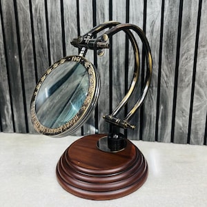 Puede incluir: Una lupa de estilo vintage con marco de latón y base de madera. La lupa tiene una lente circular y está montada sobre un brazo de latón curvado. La base es de color marrón oscuro y de forma circular.