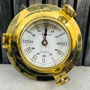 Può includere: Un orologio al quarzo rotondo color ottone con quadrante bianco e numeri romani neri. L'orologio ha un design nautico a oblò con cerniere e una lancetta dei secondi rossa. Il testo "SHTIME" e "QUARTZ" è visibile.