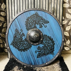 May include: A round, blue wooden shield with a black wolf design. The shield has a metal rim and a central boss. The wolf design is in a Celtic knot style. The shield is approximately 30 inches in diameter.