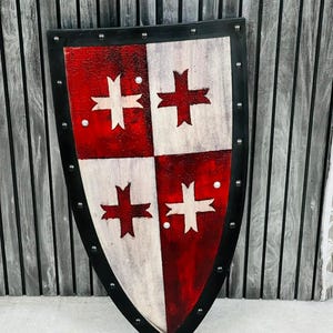 May include: A shield in a medieval style, featuring a black border with rivets. The shield's surface is split into four sections, alternating between red and white. Each section displays a white cross design. The shield has a distressed, aged appearance.