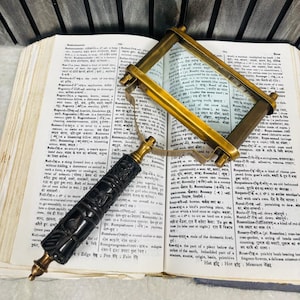 May include: A vintage brass and glass magnifying glass with a carved black handle rests on an open book. The magnifying glass has a rectangular frame. The book's pages are yellowed with text.