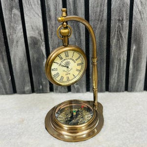 Könnte beinhalten: Eine Messing-Schreibtischuhr mit Kompassfuß. Das Zifferblatt hat römische Ziffern und lautet "Victoria London 1876". Die Uhr wird von einem gebogenen Messingständer getragen. Der Kompass ist durch eine Glasabdeckung sichtbar.