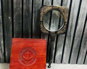 May include: A vintage magnifying glass with a square frame and a dark handle, next to a red leather case. The magnifying glass is engraved with "HENRY HUGHES SON LTD. LONDON 1941". The case has an embossed anchor logo and the text "HENRY HUGHES SONS 1941".