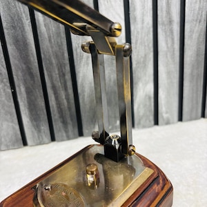 May include: Close-up of a vintage brass desk lamp with a wooden base. The lamp features adjustable arms and a small brass plate with text. The lamp's metal components have a warm, antique finish, and the base is a rich, dark wood.