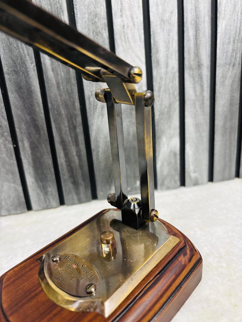 May include: Close-up of a vintage brass desk lamp with a wooden base. The lamp features adjustable arms and a small brass plate with text. The lamp's metal components have a warm, antique finish, and the base is a rich, dark wood.