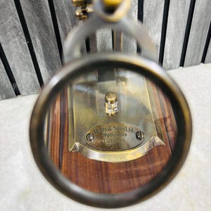 May include: Antique brass magnifying glass with a wooden base. The magnifying glass is focused on a brass plate with the inscription "Watts & Sons Ltd. Opticians London 1814". The frame is a dark bronze color.