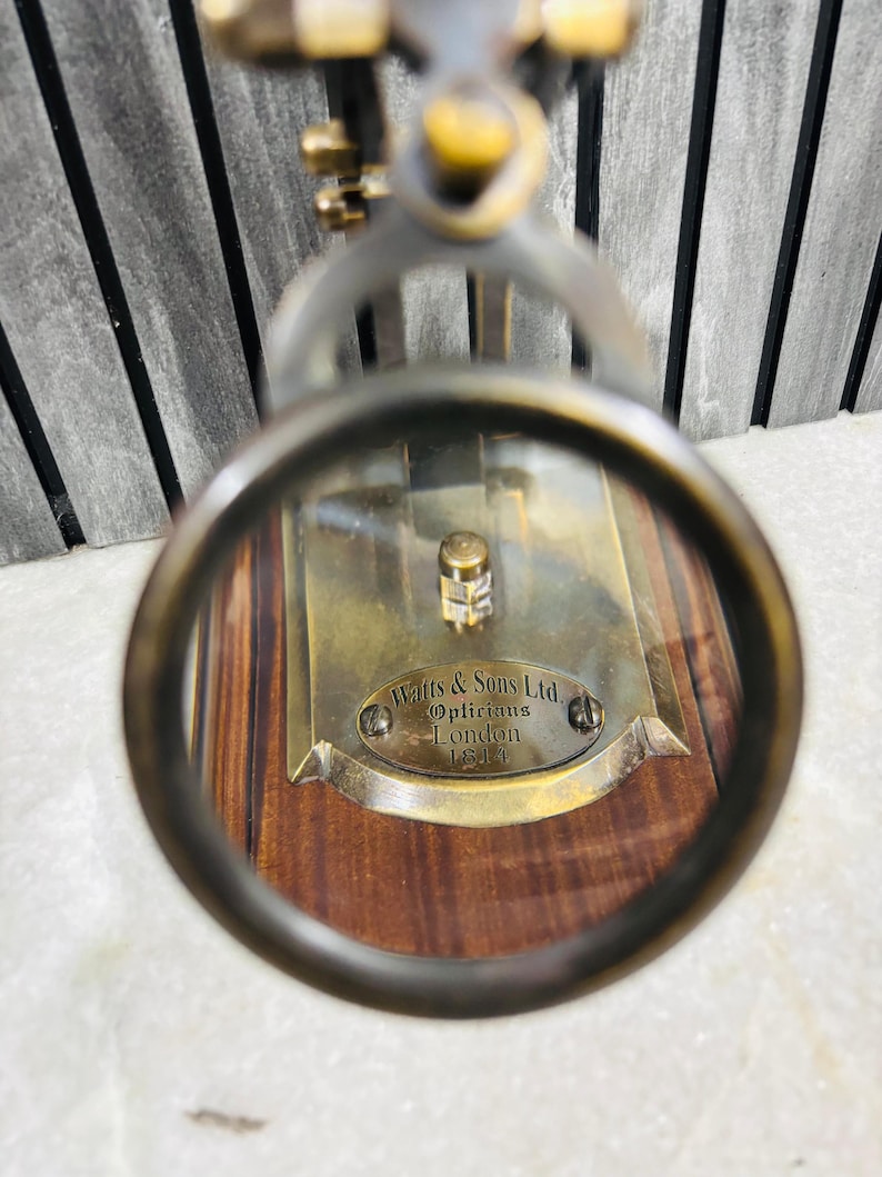 May include: Antique brass magnifying glass with a wooden base. The magnifying glass is focused on a brass plate with the inscription "Watts & Sons Ltd. Opticians London 1814". The frame is a dark bronze color.