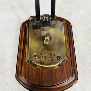 May include: A vintage optician's instrument base with a dark wood finish. The brass plate is engraved with "Watts & Sons Ltd, Opticians, London 1814". The metal arm is black and attached to the brass plate with a hinge.