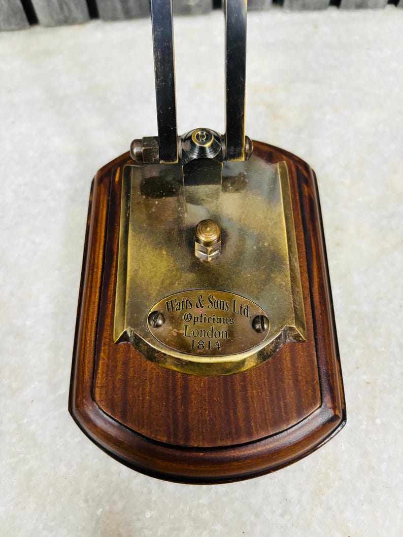 May include: A vintage optician's instrument base with a dark wood finish. The brass plate is engraved with "Watts & Sons Ltd, Opticians, London 1814". The metal arm is black and attached to the brass plate with a hinge.