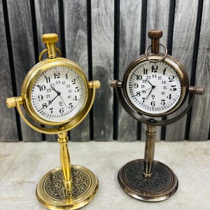 Op de afbeelding: Twee bureauklokken in antieke stijl. De ene is goudkleurig, de andere is bronskleurig. Beide hebben witte wijzerplaten met zwarte cijfers en wijzers. De klokken zijn gemonteerd op decoratieve voetstukken en hebben een lus aan de bovenkant. De wijzerplaten tonen de cijfers 1-24.