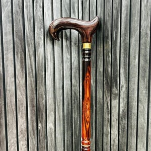 May include: A dark brown wooden walking cane with a curved handle and a decorative brass collar. The shaft of the cane shows a rich wood grain pattern. A classic walking aid, combining functionality with elegance.