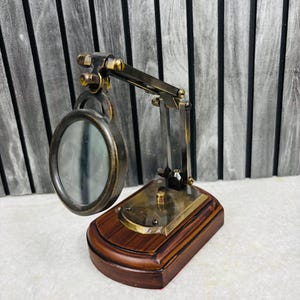 May include: A vintage-style magnifying glass with a dark brown wooden base and brass accents. The magnifying glass has a circular lens and an adjustable arm. The base is rectangular and the overall design is reminiscent of antique desk accessories.