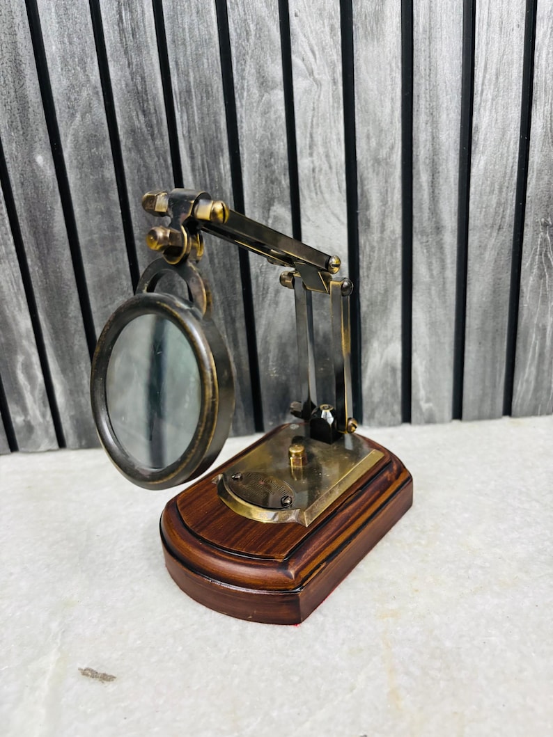 May include: A vintage-style magnifying glass with a dark brown wooden base and brass accents. The magnifying glass has a circular lens and an adjustable arm. The base is rectangular and the overall design is reminiscent of antique desk accessories.