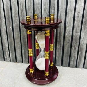 May include: A decorative hourglass with a dark red wooden frame and gold accents. The hourglass features a clear glass bulb filled with white sand. The design includes a circular base and top, connected by four red pillars with gold detailing.