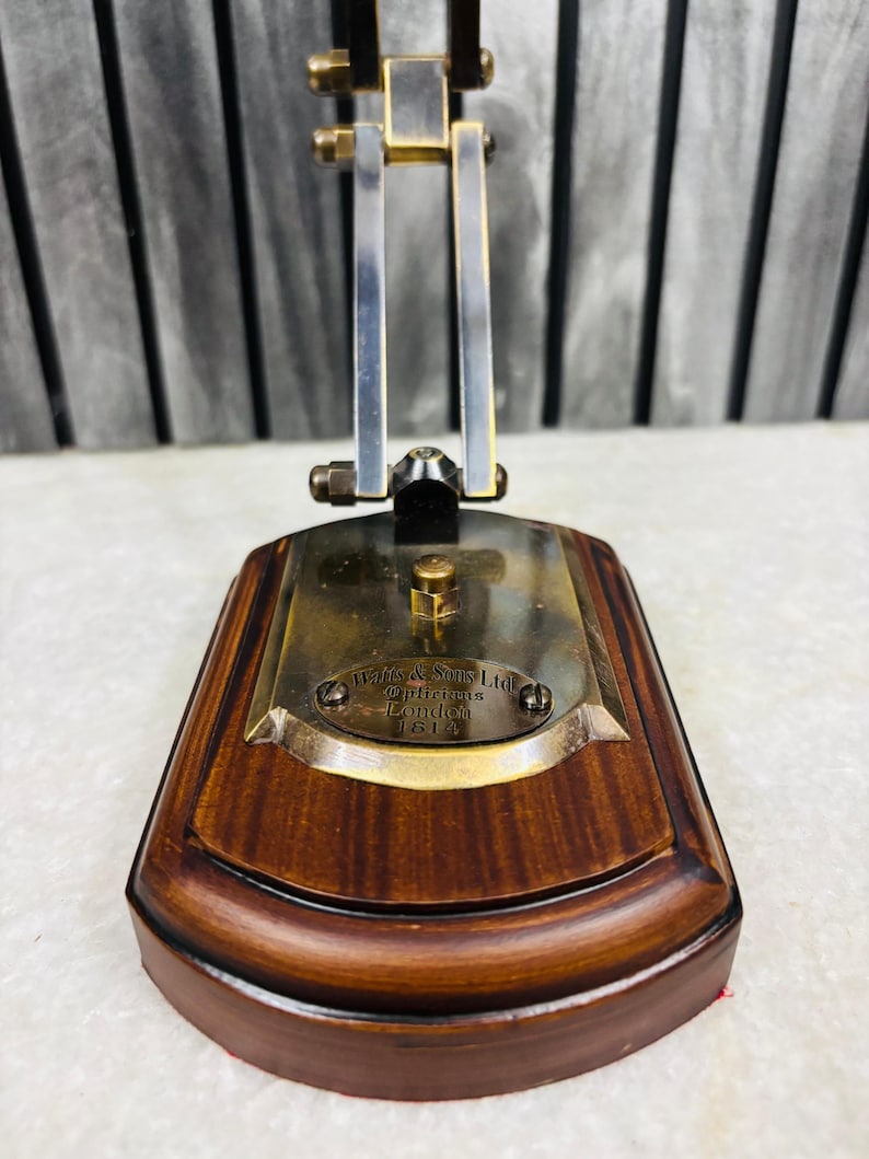 May include: A vintage brass and wood desk lamp with a rectangular base. The base is made of dark wood with a curved edge. The lamp has a brass plate with the text "W & S Sons Ltd Opticians London 1614". The lamp's frame is made of metal.