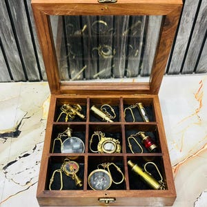 Puede incluir: Una caja de exhibición de madera con tapa de cristal, que muestra llaveros de latón en miniatura. Los llaveros incluyen brújulas, telescopios y otros artículos de temática náutica. La caja está dividida en compartimentos.