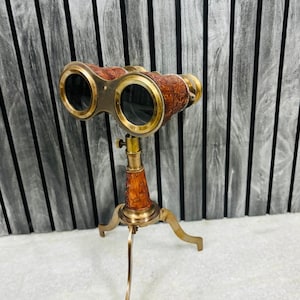 May include: Antique brass binoculars with brown leather detailing, mounted on a tripod stand. The binoculars have a classic design with a focus adjustment knob. The tripod has three curved legs, creating a vintage aesthetic.