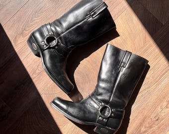 Könnte beinhalten: Ein Paar schwarze Motorradstiefel aus Leder mit eckiger Spitze und einem mittelhohen Schaft. Die Stiefel haben einen Riemen mit silberfarbenem Ring und Schnalle. Die Stiefel werden auf einer Holzoberfläche präsentiert.