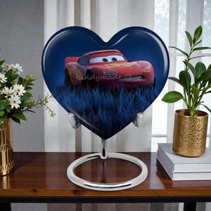 May include: A heart-shaped decorative piece featuring a red cartoon race car against a blue background. The car is set in a field of blue grass. The piece is supported by a silver metal stand.