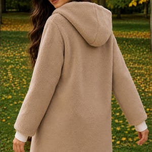 May include: A beige hooded jacket made of textured fleece material. The jacket features long sleeves with white ribbed cuffs and a hood. The design is simple and functional, providing a soft, fuzzy appearance with a consistent light brown color.